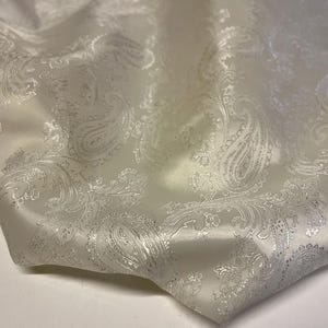 Può includere: Tessuto bianco sporco con un motivo paisley argentato. Il tessuto è piegato, mostrando la consistenza e la lucentezza del materiale. Il motivo paisley è dettagliato e copre l'intera superficie.