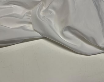 May include: A close-up of white fabric with a silky, flowing texture. The fabric is draped and folded, creating soft shadows and highlights. The material appears to be lightweight and possibly used for clothing or decorative purposes.
