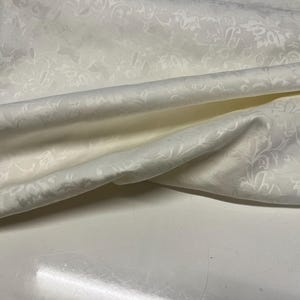 May include: A length of white fabric featuring a tonal damask pattern with intricate floral and leaf motifs. The material has a subtle sheen and is draped to show soft folds and curves, highlighting the texture and the repeating woven design.