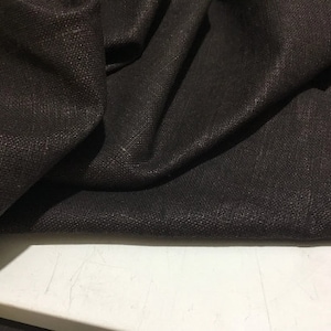 Puede incluir: Primer plano de una tela marrón oscura con tejido texturizado. La tela está doblada y drapeada sobre una superficie blanca.