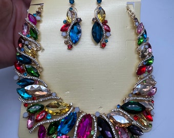 Könnte beinhalten: Ein farbenfrohes Statement-Halsketten- und Ohrring-Set mit einer goldenen Basis und mehrfarbigen Strasssteinen. Die Halskette hat ein kaskadierendes Design mit verschiedenen Formen und Größen von Strasssteinen. Die Ohrringe sind im Hänge-Stil mit einem ähnlichen Design wie die Halskette. Die Karte lautet "Fashion Jewelry".