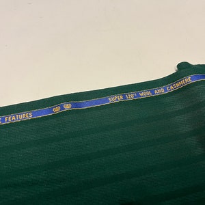 May include: Close-up of a dark green fabric with a blue and gold ribbon that reads "SUPER 120's WOOL AND CASHMERE" and "THE ELEGANCE FEATURES".