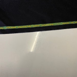Op de afbeelding: Close-up van een zwarte stof met een groen en gouden label met de tekst "SUPERFINE WOOL WORSTED FABRIC 100% WOOL"