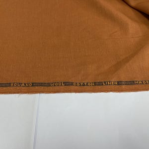 Op de afbeelding: Een close-up van een bruine stof met de tekst "SOLANO WOOL COTTON LINEN MADE IN ENGL" gedrukt op een donkerbruine strook. De stof heeft een effen, warme bruine kleur.