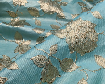 May include: A light blue fabric with a gold floral pattern. The fabric has a subtle sheen and a textured surface.