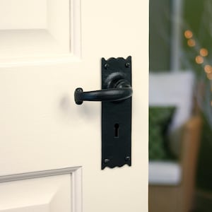 May include: A black door handle with a keyhole and an ornate design. The handle is fitted to a white door.