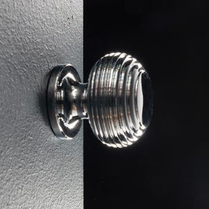 May include: A chrome cabinet knob with a round, ridged design. The knob has a smooth, white centre.