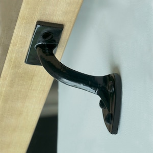 May include: Black metal bracket with a curved arm and a flat base. The bracket is attached to a wooden surface.