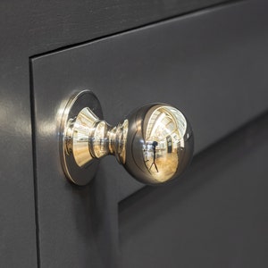 May include: A polished silver door knob with a round, reflective handle. The knob is mounted on a dark grey door.