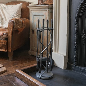 May include: A black metal fireplace tool set with a stand, in front of a black fireplace with a fire burning inside. The tools are in a spiral design and include a poker, tongs, and shovel. The fireplace is in a room with a leather armchair and a wooden table.