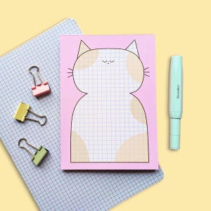 Op de afbeelding: Een roze notitieblok in de vorm van een kat met een rasterpatroon, samen met een lichtblauwe pen. Drie paperclips in roze, geel en groen zijn ook aanwezig. Het notitieblok ligt op een blauw geruit papier, tegen een gele achtergrond.