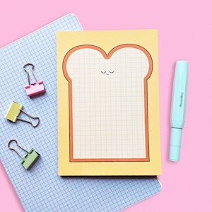 May include: A yellow notepad shaped like a slice of bread with a smiling face, orange outline, and grid pattern. A light blue pen and three binder clips in pink, yellow, and green are also visible on a blue grid notebook, all against a pink background.