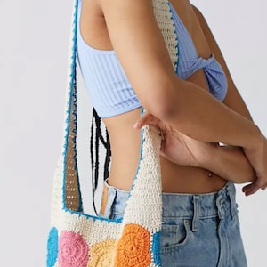 May include: A white crochet tote bag with a colorful geometric pattern. The bag has a long strap and is made of a soft, lightweight material.