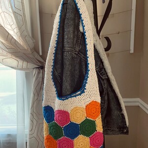 May include: A white crocheted tote bag with a colorful geometric pattern of hexagons in shades of pink, orange, yellow, green, and blue. The bag is hanging on a black metal hook.