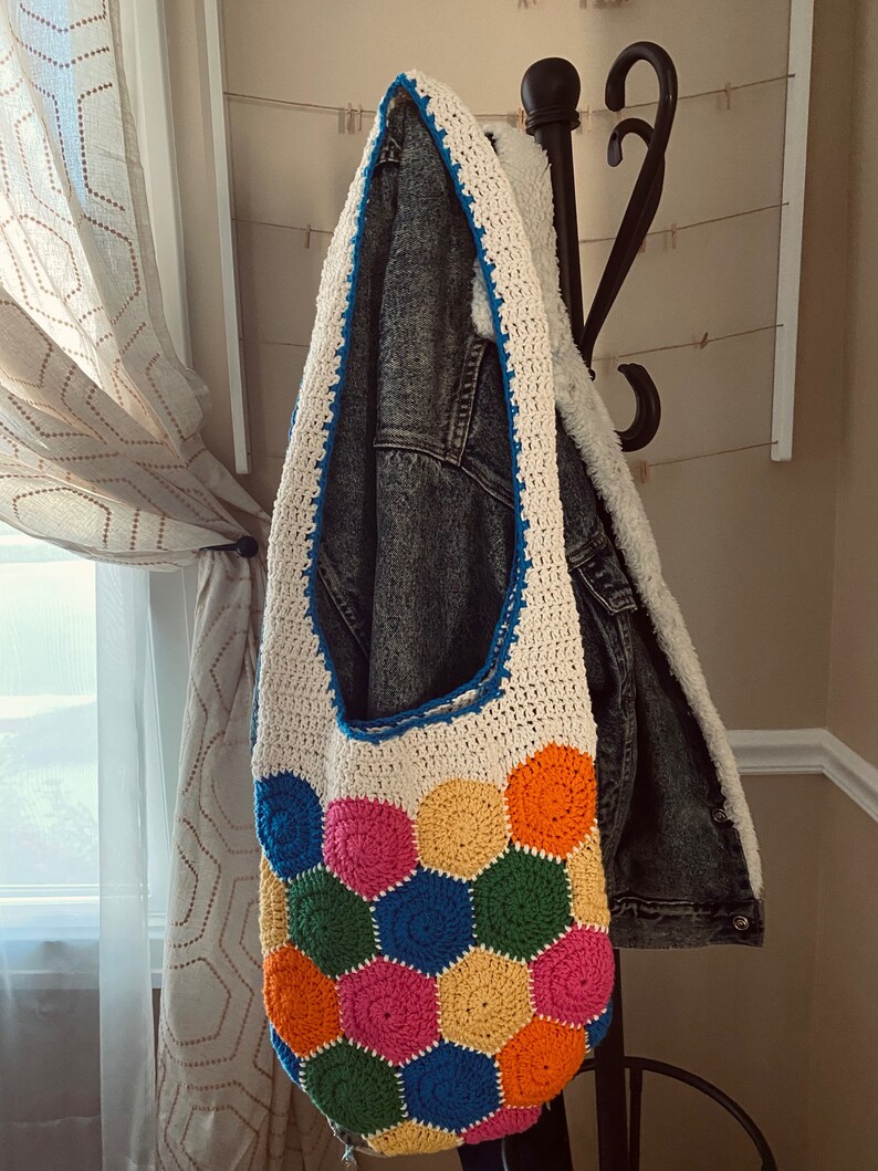 May include: A white crocheted tote bag with a colorful geometric pattern of hexagons in shades of pink, orange, yellow, green, and blue. The bag is hanging on a black metal hook.