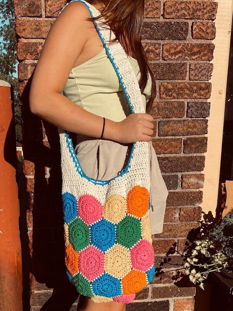 May include: A white crocheted tote bag with a colorful geometric pattern. The bag has a long strap and is made of thick yarn. The colors of the pattern are pink, orange, yellow, green, and blue.