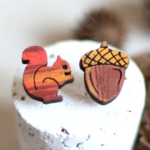 Peut inclure: Une paire de boucles d'oreilles en bois représentant un écureuil rouge et orange et un gland brun avec un chapeau jaune.
