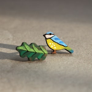 May include: A pair of wooden earrings featuring a bluebird and an oak leaf. The bluebird is painted yellow with blue wings and white spots. The oak leaf is painted green.