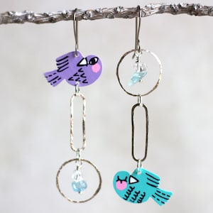Op de afbeelding: Een paar grillige oorhangers. Eén oorbel heeft een paarse vogel, de andere een turquoise vogel, beide met zwarte details en roze accenten. De vogels hangen aan zilverkleurige metalen hoepels en langwerpige schakels, met kleine blauwe kristallen kralen.