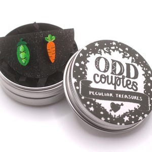 Könnte beinhalten: Eine silberne Dose mit den Worten "ODD couples PECULIAR TREASURES" auf dem Deckel. Im Inneren befindet sich ein schwarzes Filzstück mit einer grünen Erbsenschote und einer orangefarbenen Karotte. Die Erbsenschote hat drei grüne Erbsen, und die Karotte hat grüne Blätter.