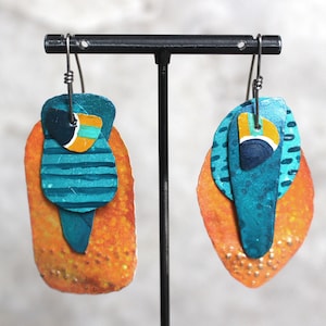 May include: A pair of dangle earrings with a layered design. The earrings are made of metal and painted in shades of orange, teal, and blue. The earrings are suspended from silver hooks.