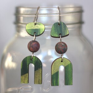 May include: A pair of green and gold geometric earrings with a red and brown bead accent. The earrings are made of wood and have a modern design.