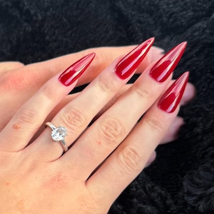 Pode incluir: Uma mão com unhas longas, pontudas e vermelhas e um anel de prata com uma pedra clara e redonda. A mão repousa sobre uma superfície preta e texturizada.