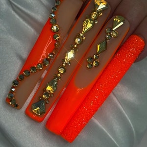 May include: Long, bright orange acrylic nails with a nude base and gold glitter accents. Some nails have a sparkly orange finish.