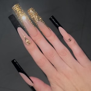 May include: Long, nude-colored acrylic nails with black tips and gold glitter accents. Some nails have small gold rhinestones.