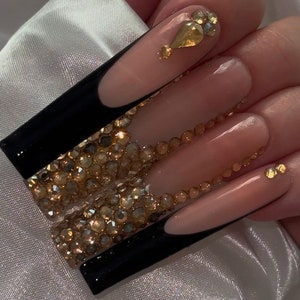 May include: Long, nude-colored acrylic nails with black tips and gold rhinestones.
