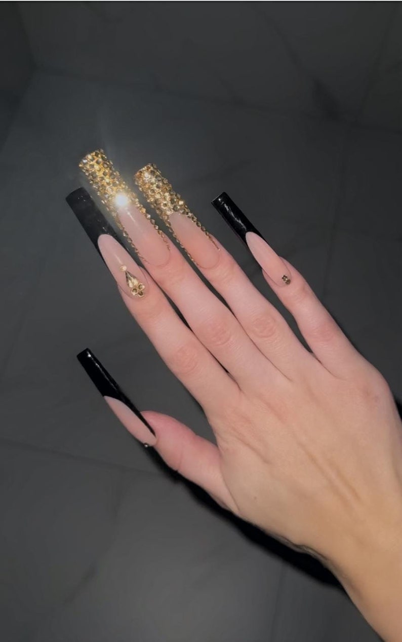 May include: Long, nude-colored acrylic nails with black tips and gold glitter accents. Some nails have small gold rhinestones.