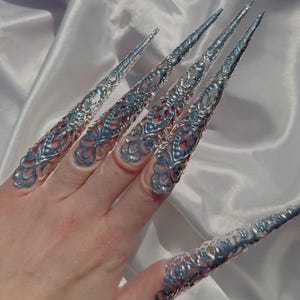 Puede incluir: Anillos de uñas de filigrana plateada con puntas puntiagudas. Los anillos se llevan en varios dedos y están diseñados para parecerse a uñas largas y afiladas.