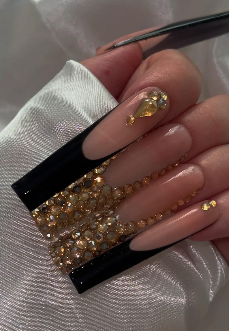 May include: Long, nude-colored acrylic nails with black tips and gold rhinestones.