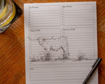 May include: A white notepad with sections for Top Priorities, Appointments, Today's To-Do, Tomorrow To-Do, and Notes. A pen rests on the notepad. A sketch of a longhorn is in the center. The notepad is on a wooden surface.