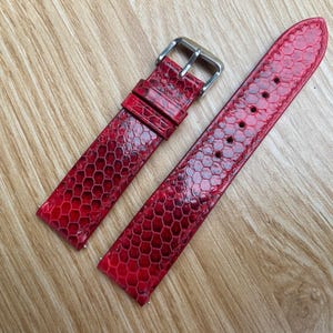 Puede incluir: Dos correas de reloj rojas con un patrón texturizado similar a la piel de reptil. Las correas tienen un acabado brillante y una hebilla plateada. Se muestran sobre una superficie de madera marrón claro. Miden unos 20 cm.