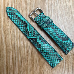 May include: A pair of turquoise and black snakeskin-patterned watch straps. The straps are made of leather and feature a silver-colored buckle. The watch bands are displayed on a light brown wooden surface.