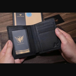 Puede incluir: Cartera de cuero negro con múltiples ranuras para tarjetas y una ventana de identificación transparente. La cartera presenta un emblema dorado y el texto "ALPHA LEATHER". El interior es negro con costuras blancas. La cartera está abierta.