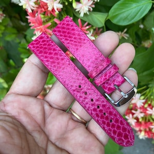 May include: Bright pink watch straps with a snakeskin texture. The straps have a silver-colored buckle and several holes for adjustment. The background features green foliage and colorful flowers.