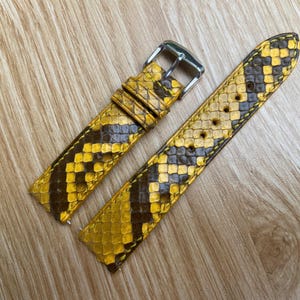 Puede incluir: Una correa de reloj de cuero con estampado de serpiente amarillo y marrón. La correa tiene una hebilla plateada y está diseñada para sujetarse a un reloj. El cuero tiene un aspecto texturizado.