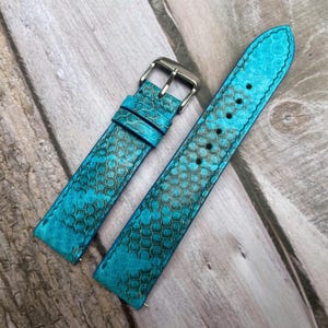Puede incluir: Correa de reloj turquesa de dos piezas con estampado de piel de serpiente. La correa tiene una hebilla plateada y está hecha de un material texturizado. La correa del reloj se muestra sobre una superficie de madera.