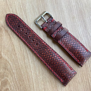 May include: A brown leather watch strap with red stitching. The strap has a textured surface and is designed for a wristwatch.