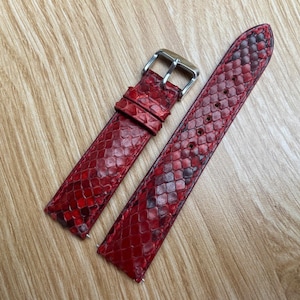 Puede incluir: Correa de reloj roja con un patrón de piel de serpiente. La correa es de cuero y tiene una hebilla plateada. La correa del reloj es de color rojo vibrante con detalles en rojo oscuro y negro.