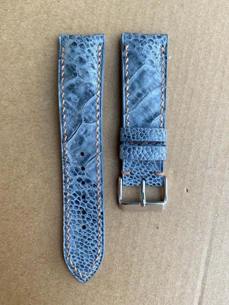 May include: A gray leather watch strap with a light brown stitching and a silver buckle.