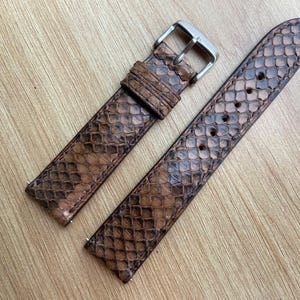 May include: A two-piece watch strap featuring a brown and black snakeskin pattern. The strap includes a silver-toned metal buckle and a matching sliding keeper loop. The texture displays a distinct scale design across the entire surface of the band.
