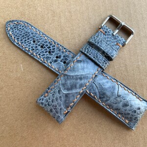 May include: A gray leather watch strap with a silver buckle and orange stitching. The strap is made of a textured material that resembles alligator skin.