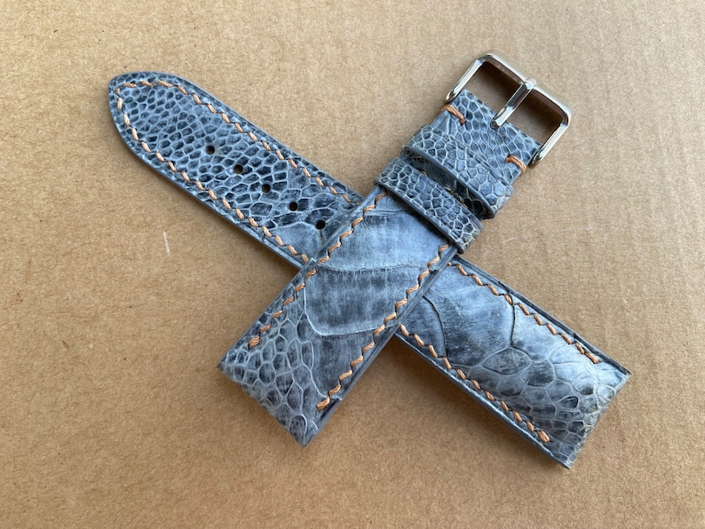 May include: A gray leather watch strap with a silver buckle and orange stitching. The strap is made of a textured material that resembles alligator skin.