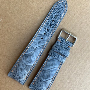 May include: A gray ostrich leather watch strap with brown stitching. The strap is shown in two pieces, one with a buckle and one without.