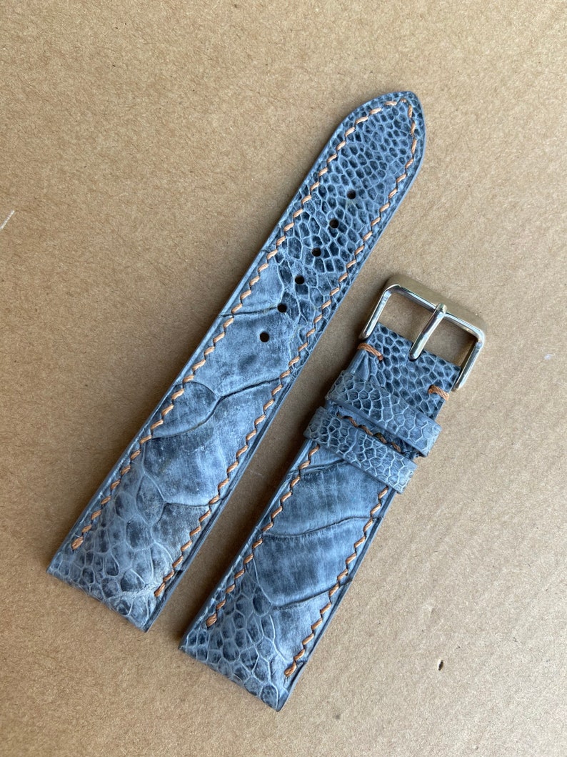May include: A gray ostrich leather watch strap with brown stitching. The strap is shown in two pieces, one with a buckle and one without.
