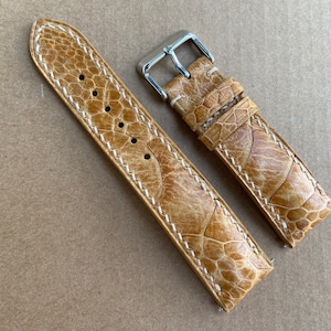 Könnte beinhalten: Ein hellbraunes Lederarmband mit weißen Nähten. Das Armband ist aus einem strukturierten Material, das Krokodilleder ähnelt.