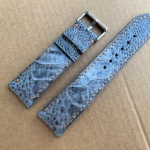 May include: A gray ostrich leather watch strap with brown stitching and a silver buckle.